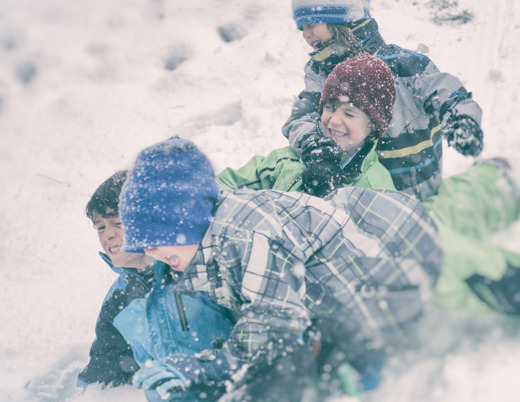 A group of four boys in blue and green snow gear wrestle in the snow.