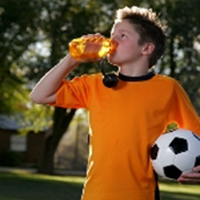A boy in an orange jersey holding a soccer ball and drinking from a water bottle.
