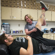 An athletic trainer stretches a teenage athlete's leg while he lies on a bench.
