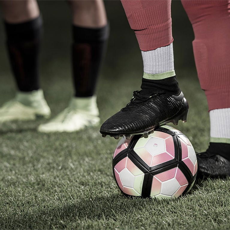 A closeup of a teenage athlete's feet, with one on top of a soccer ball on the field.