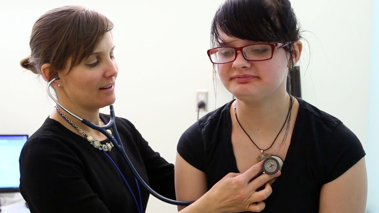 A woman in a black top with her hair pulled back in a bun and a stethoscope on listens to the heart of a teen girl wearing a black T-shirt and red glasses with her hair in a ponytail.  