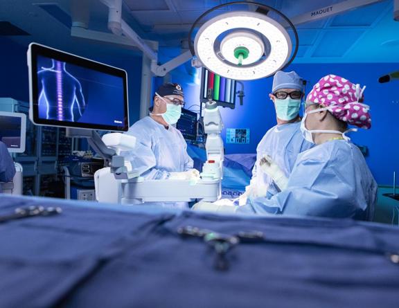 Three doctors in scrubs and masks stand in the operating room around a white surgical robot. There is a screen with a digital image of a patient’s spine.