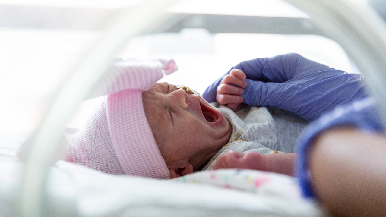 Infant with mouth open, pink hat and blue blanket in the NICU. 