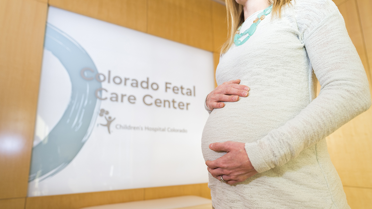 A pregnant parent holds their belly while standing in front of a sign for the Colorado Fetal Care Center.