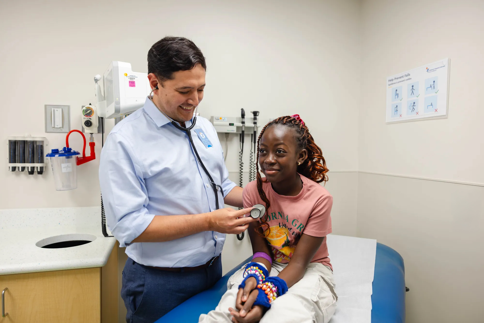 A doctor uses a stethoscope on a pediatric patient sitting on an exam table and smiling.