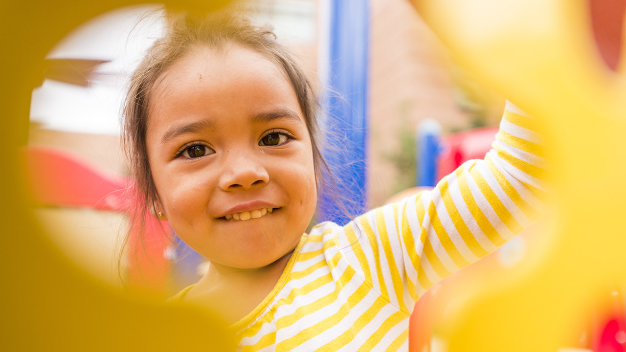 A young girl in a yellow and white striped shirt plays in a jungle gym.