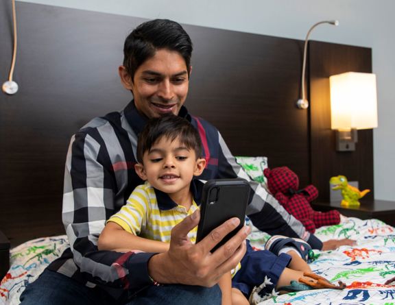 A parent and child sit in bed and look at the screen of a phone in the adult’s hand.